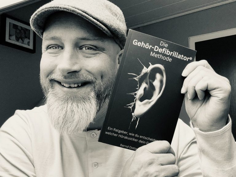 Mann mit Bart und Mütze, der ein Buch über Hörgesundheit hält und lächelt.