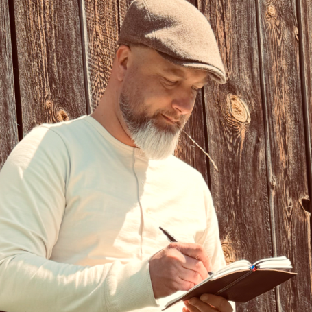 Mann mit Hut schreibt in ein Notizbuch vor einer Holzwand.