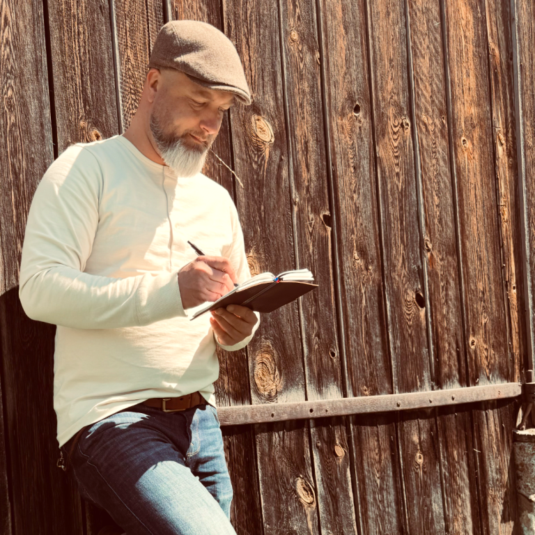 Ein Mann mit Hut steht an einer Holzwand und macht Notizen in einem Notizbuch.